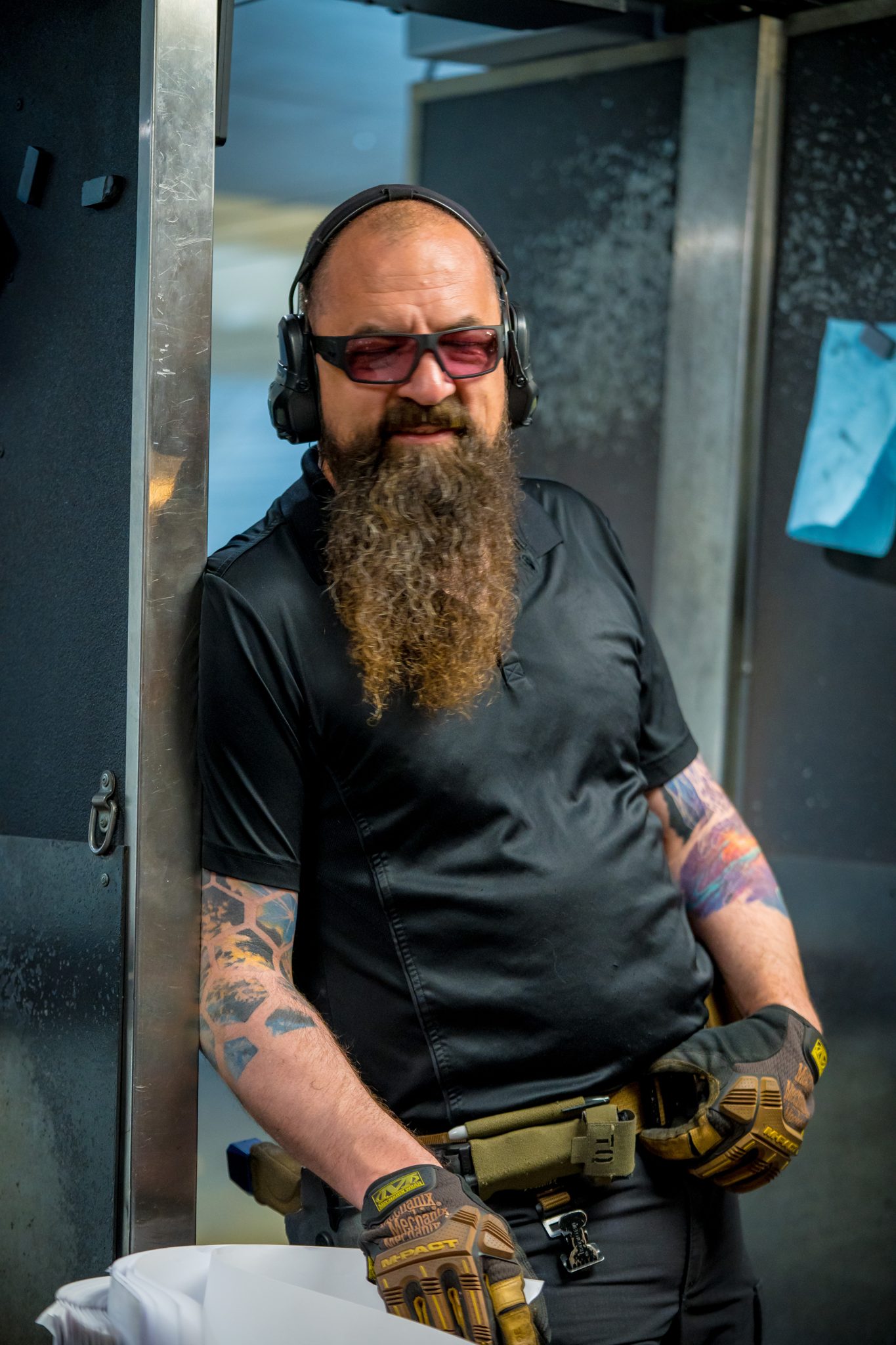 A bearded Machine Guns Vegas Range Safety Officer in ear and eye protection, gloves and a holster, standing in an indoor lane
